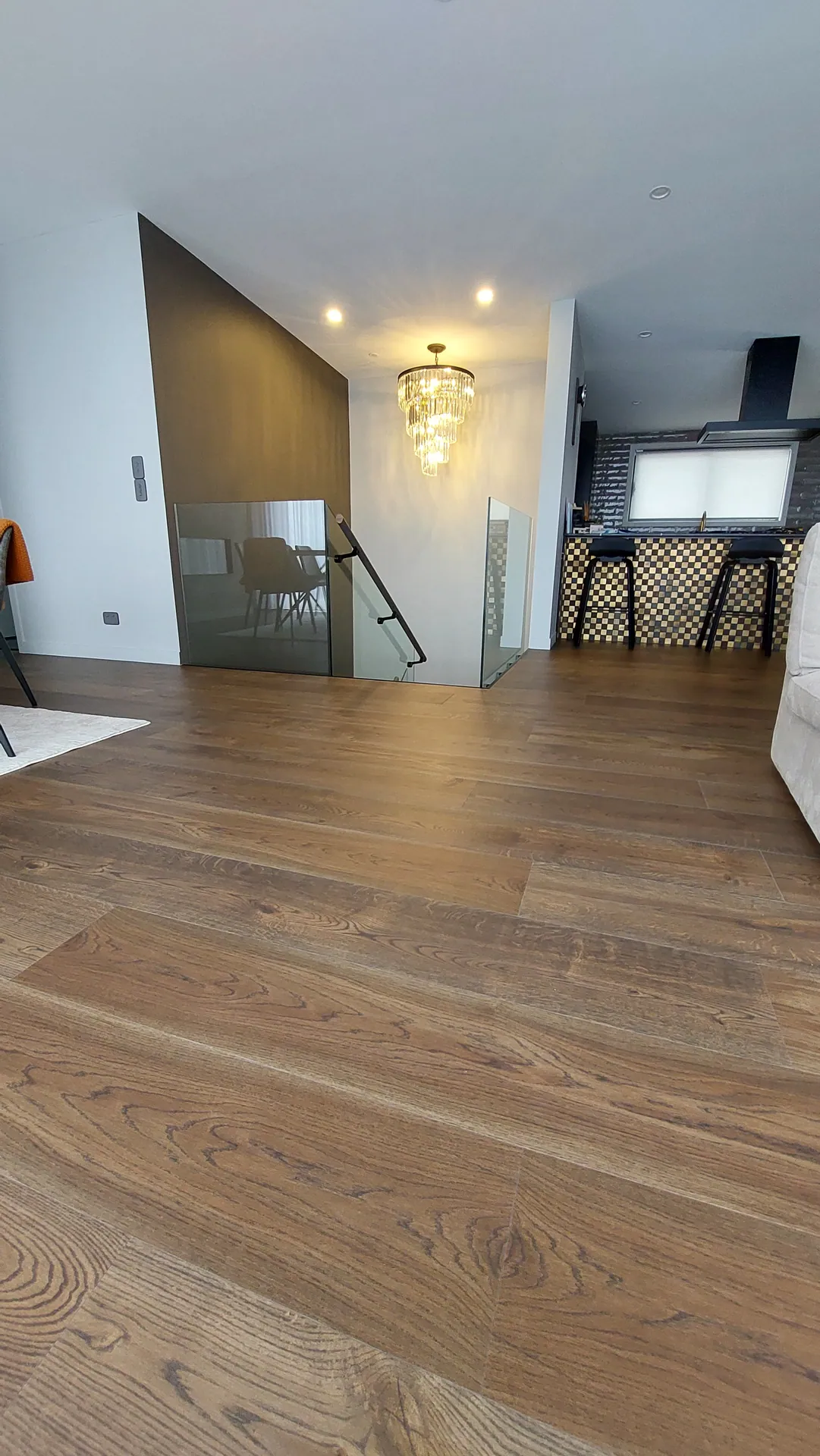 Wide brown oak boards in a living area beside a glass stair balustrade, with a chandelier and kitchen stools behind.
