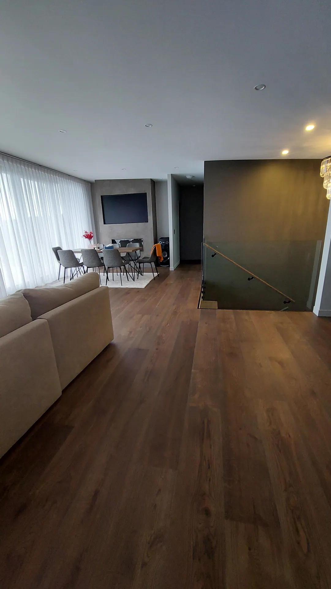 A lounge and dining area with a wide brown oak floor, a beige sofa and a glass stair rail.