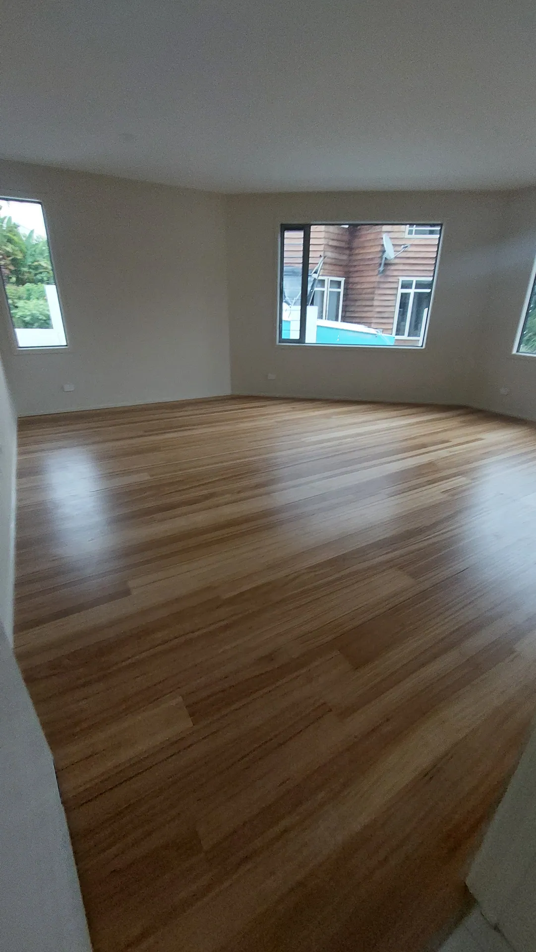 An empty room with a glossy honey-toned strip timber floor and windows on two walls.