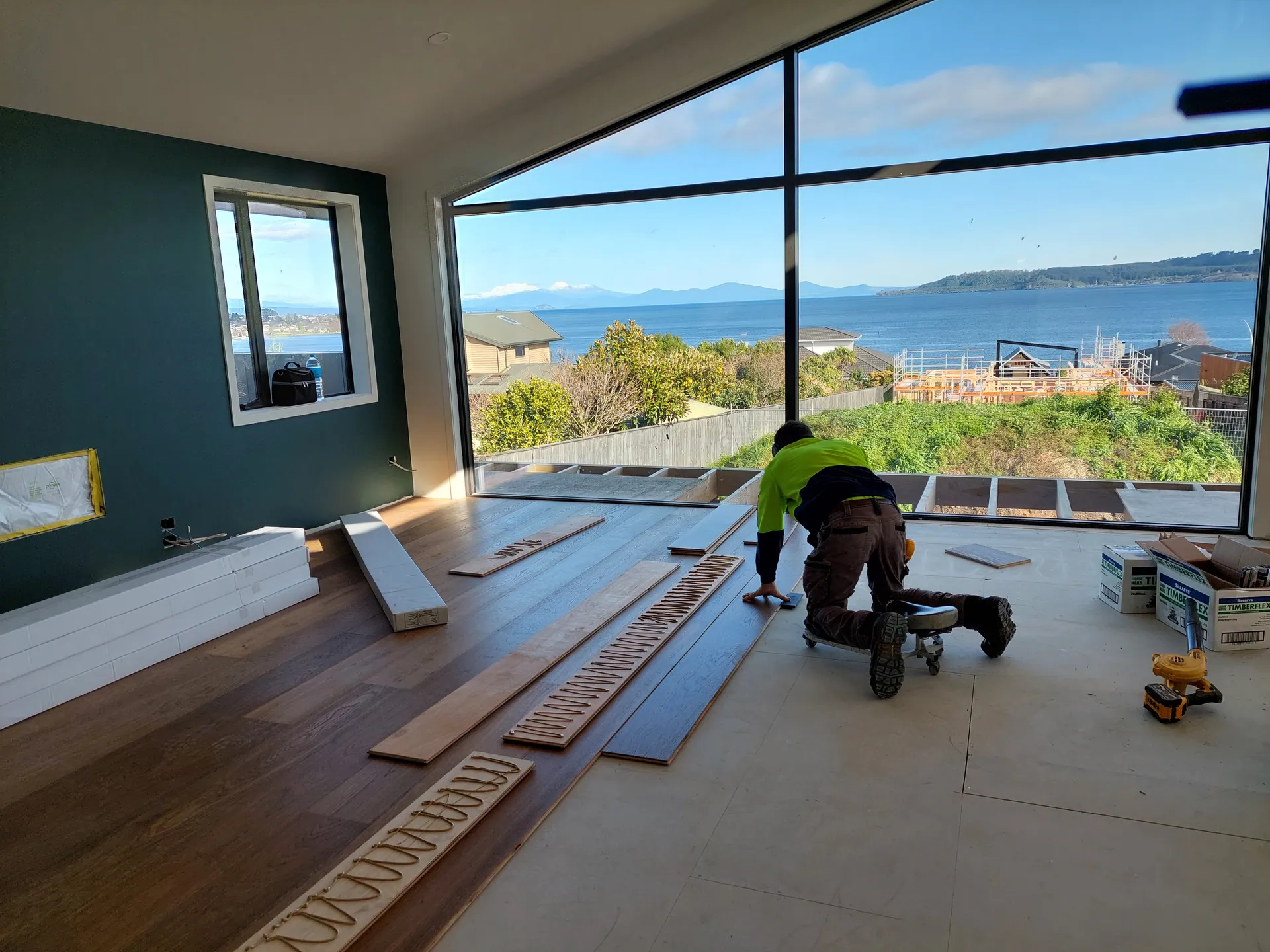 Floor layer kneeling on a creeper laying dark timber boards in a room with a large window over the sea.