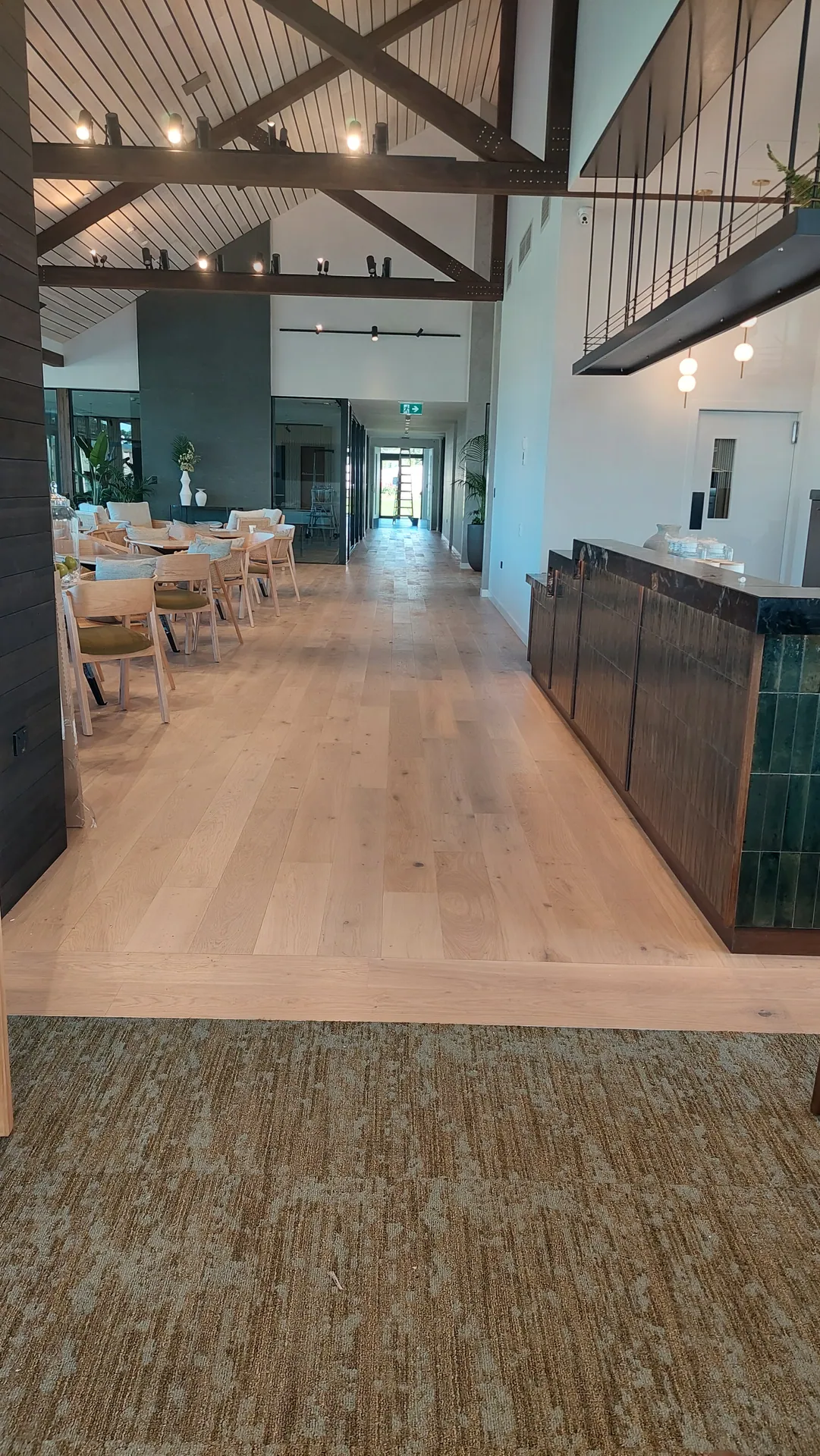 A wide light oak floor runs past a dark bar counter and dining tables under exposed roof trusses, with carpet tiles in the foreground.