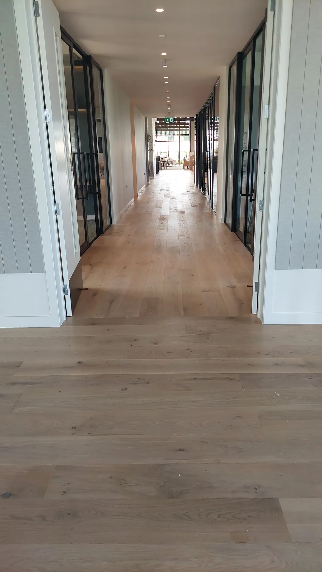 A long corridor with light oak boards between glass-panelled doors, leading to a bright room at the far end.