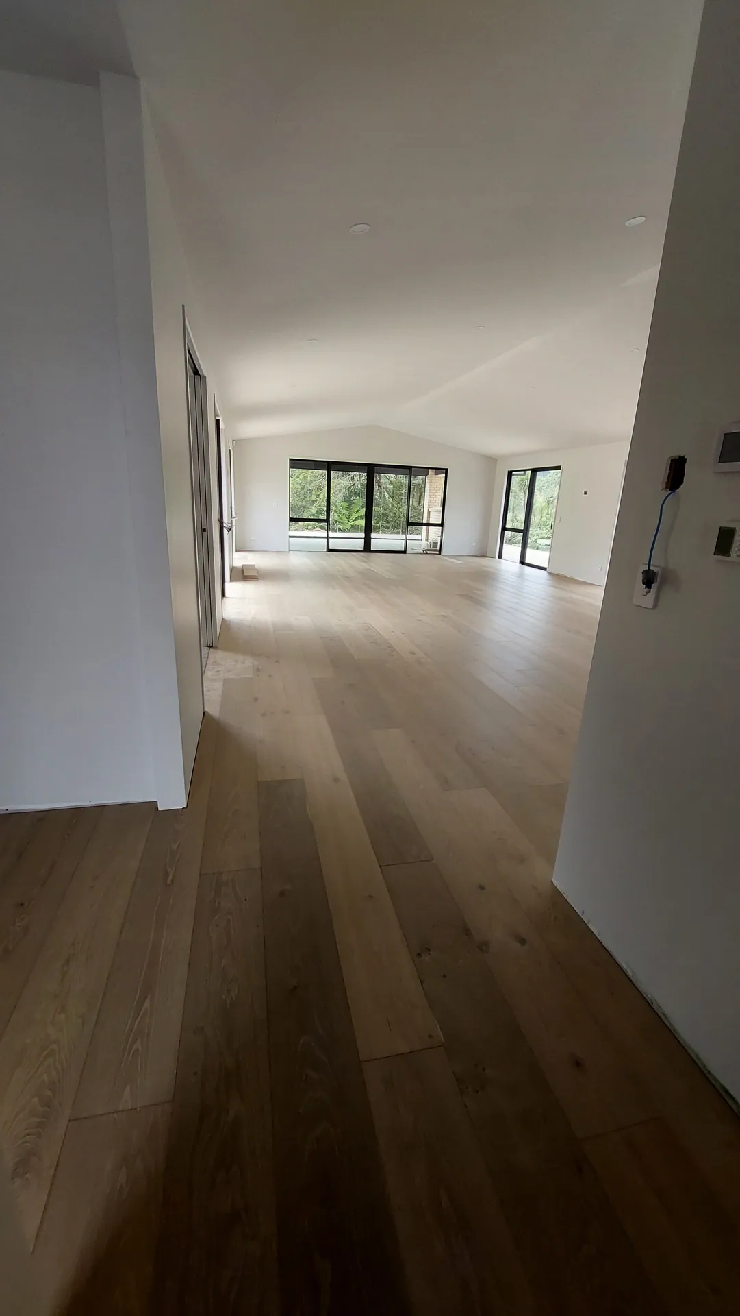 A long hallway with wide oak boards leading into a bright living area with large windows.
