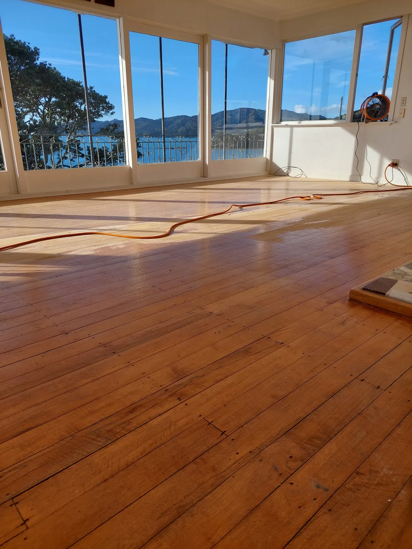 Sanded native timber floor partly coated in a sunny room with windows over a harbour, an extension cord across the floor.
