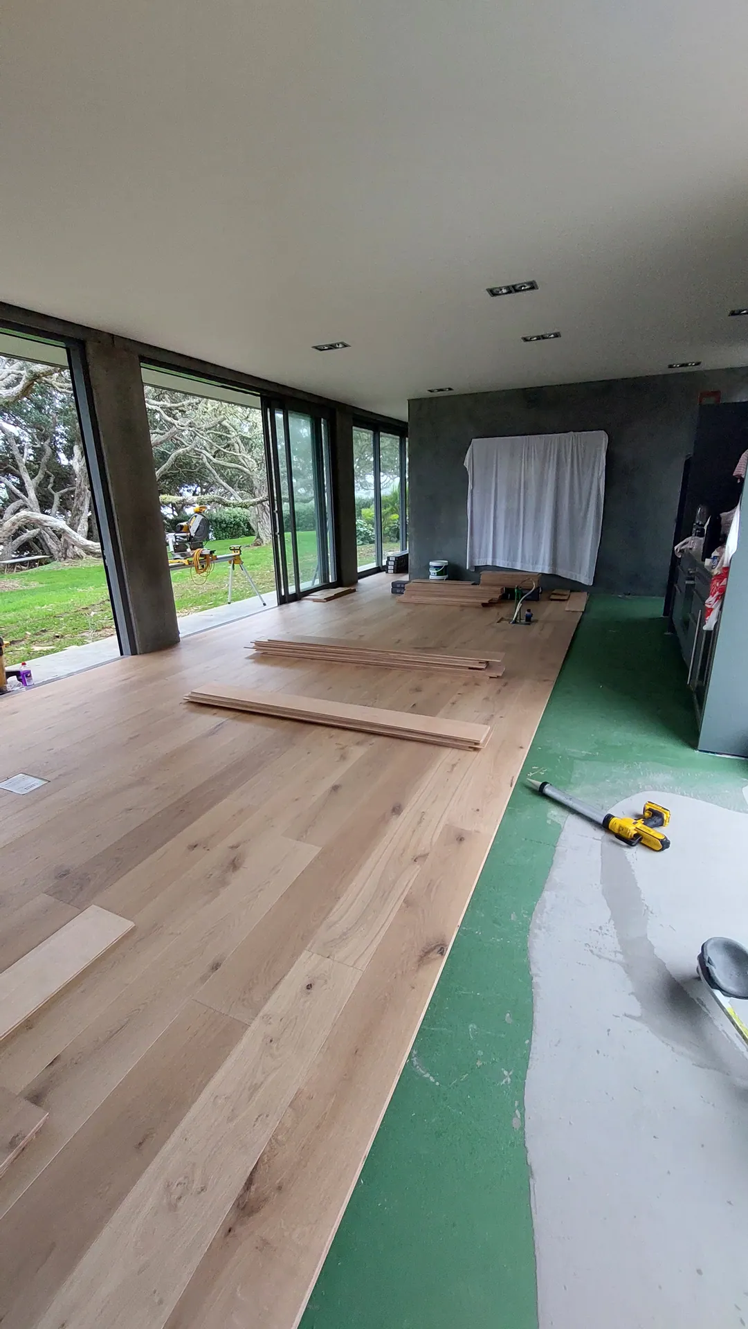 Light oak boards partly laid over a green membrane in a glass-walled room, with loose boards and a drill on the floor.