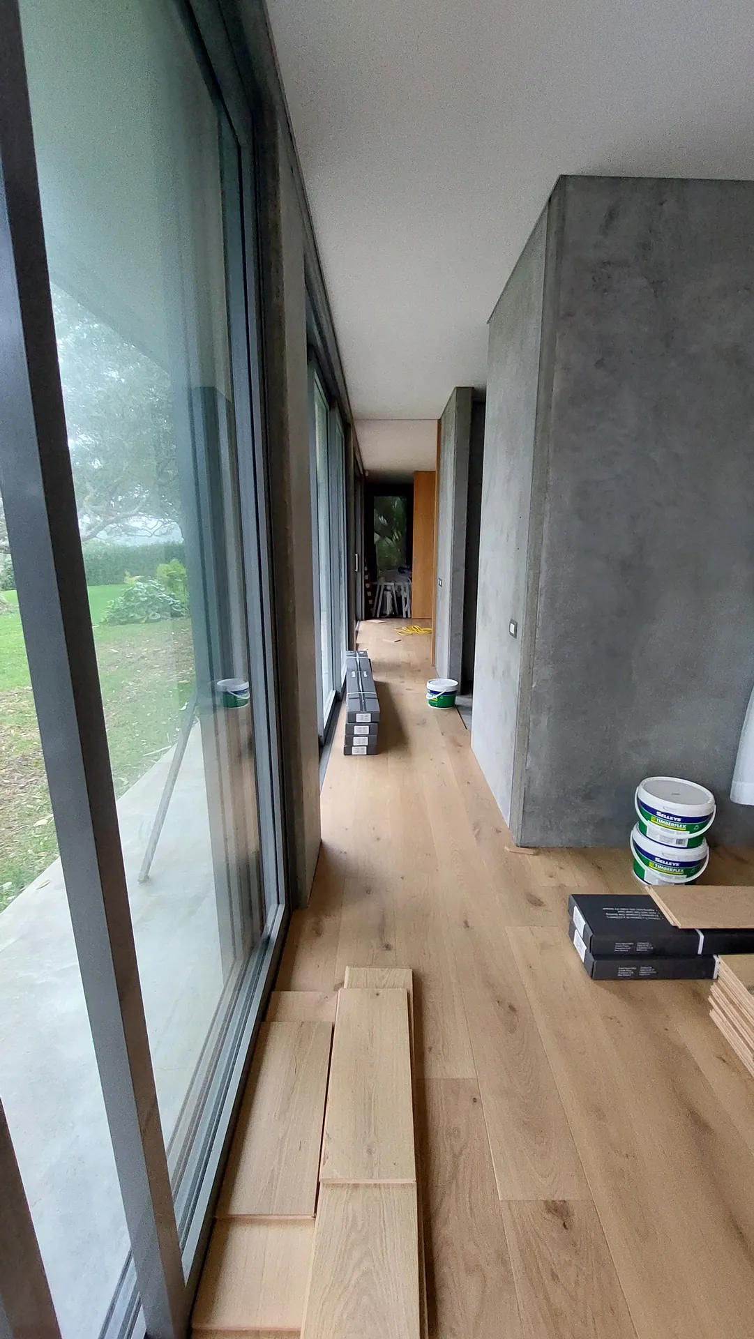 A narrow strip of light oak boards being laid along a row of glass doors, with loose boards and adhesive tubs beside.