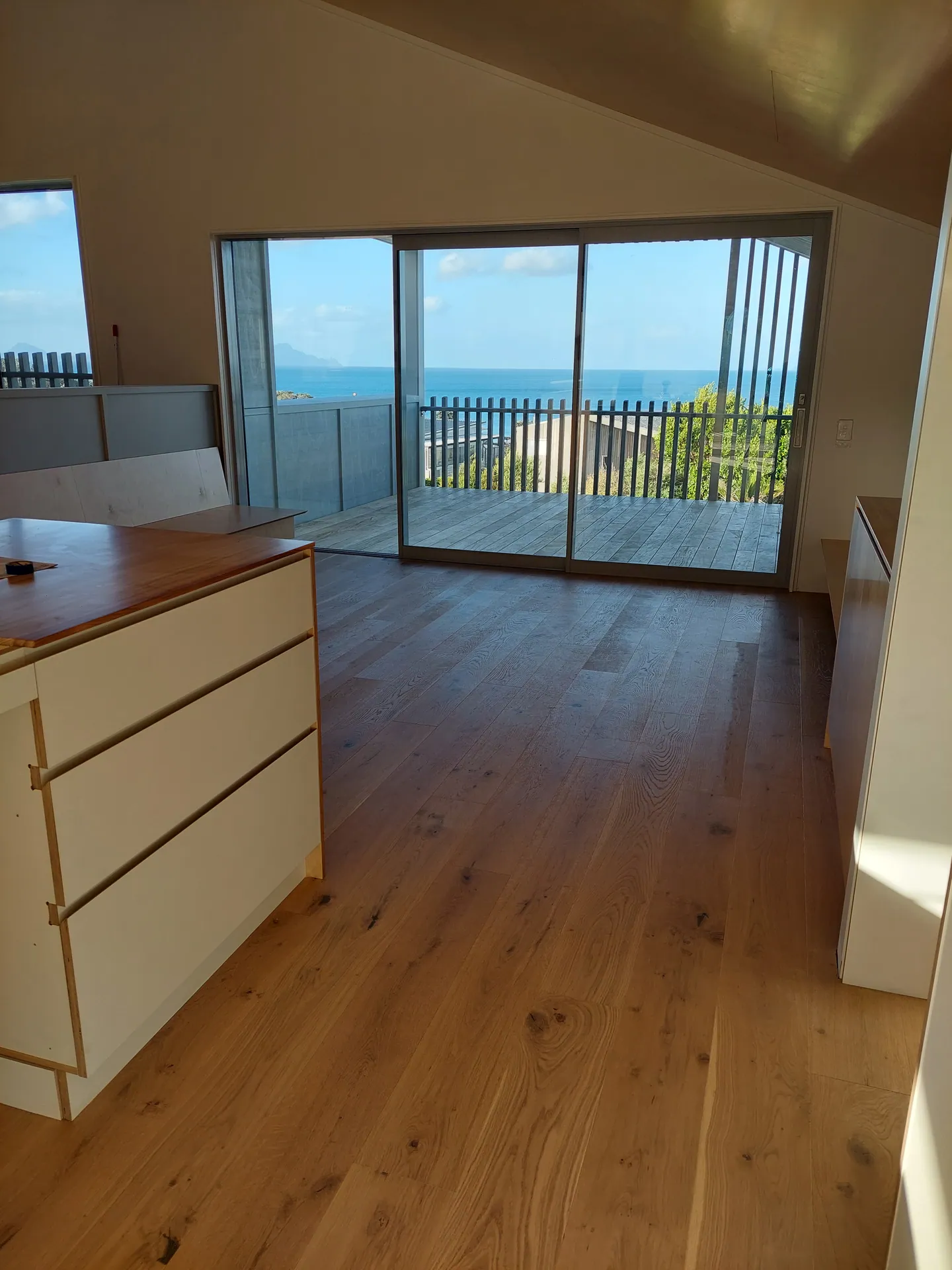 Wide oak floor running past a kitchen island to sliding doors and a deck with a sea view.