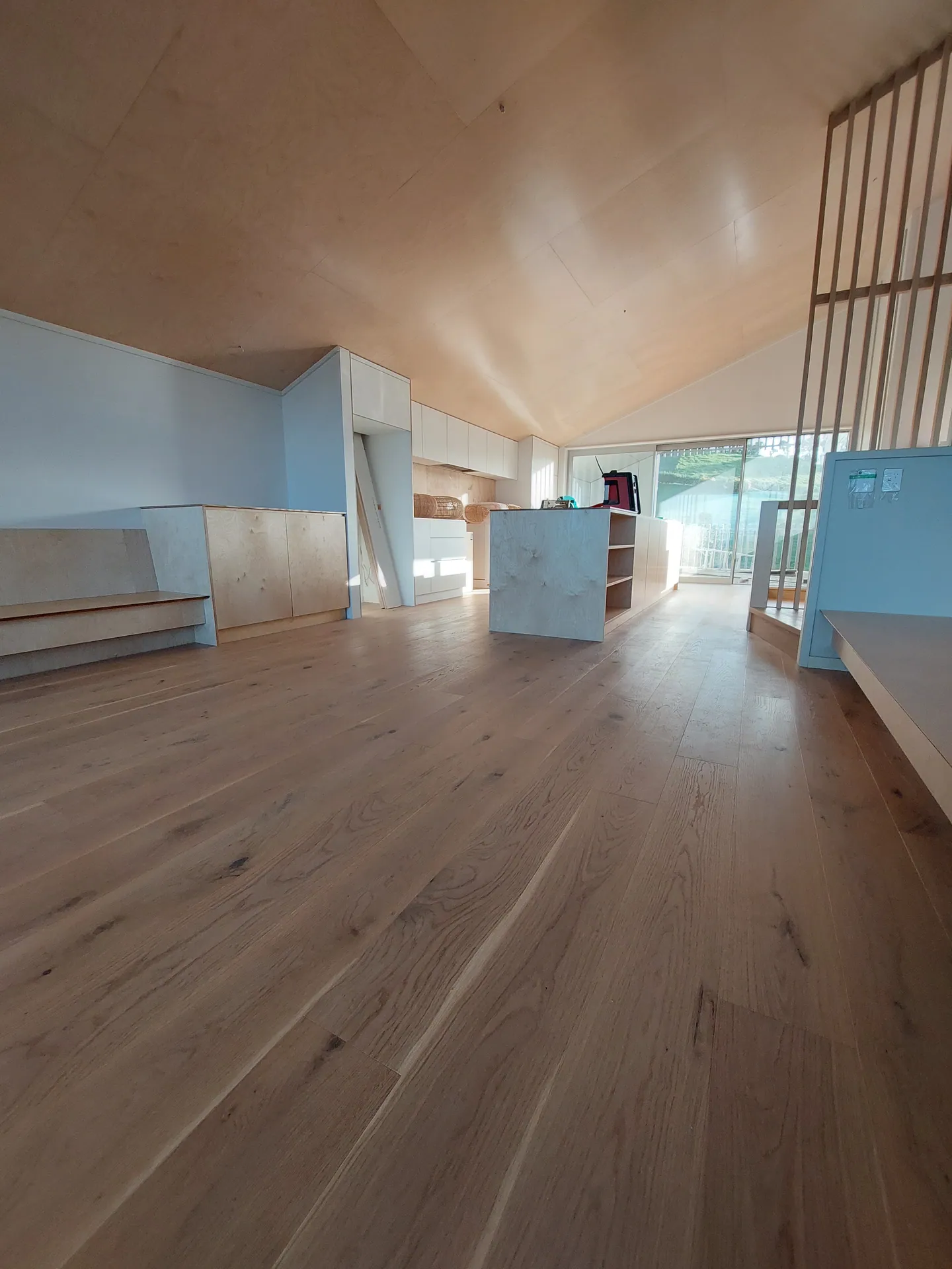 Oak floor running through an open-plan living and kitchen area under a plywood ceiling.