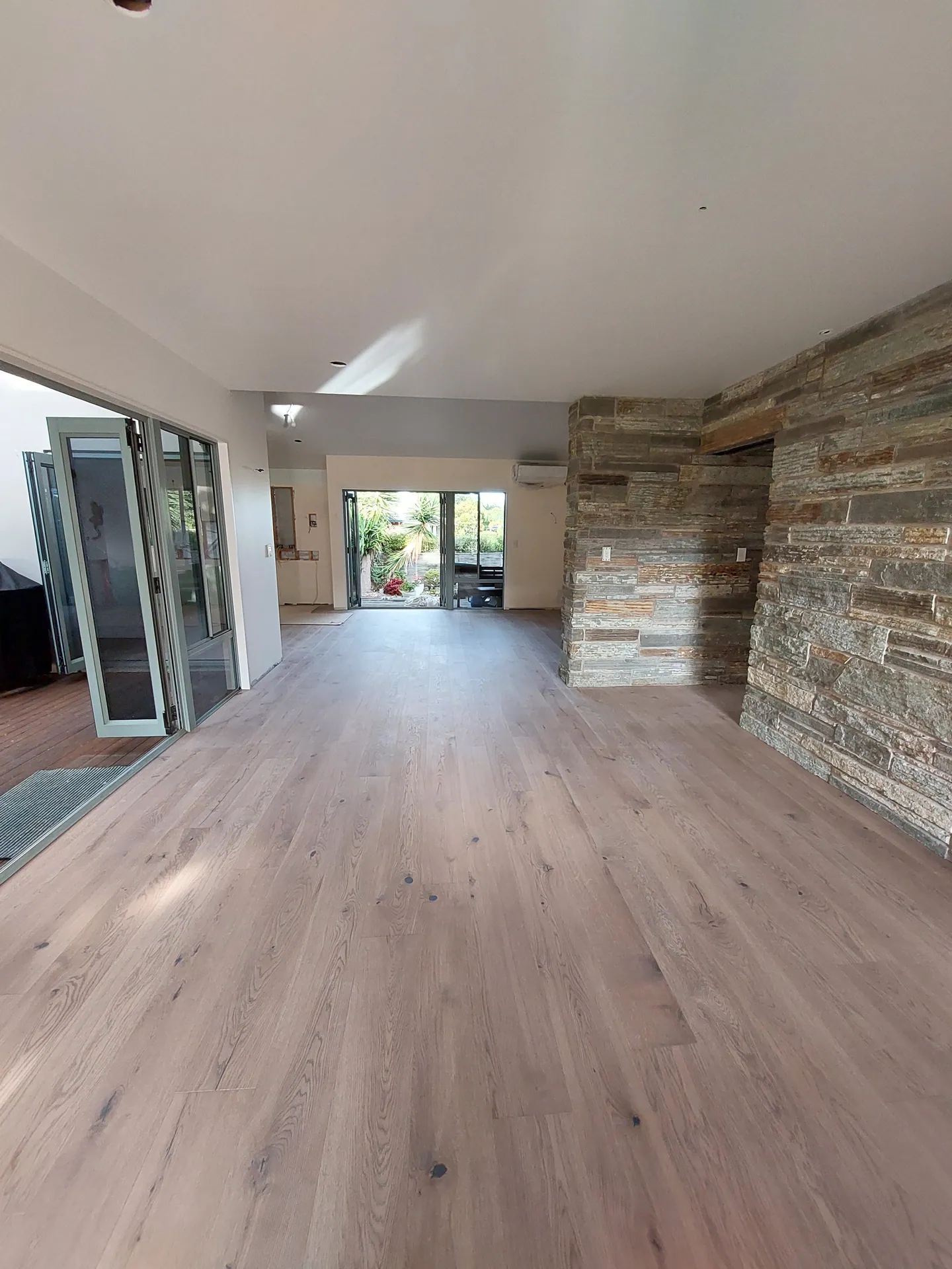 Long open living space with grey-toned wide plank timber floor, a stone feature wall and bifold doors.