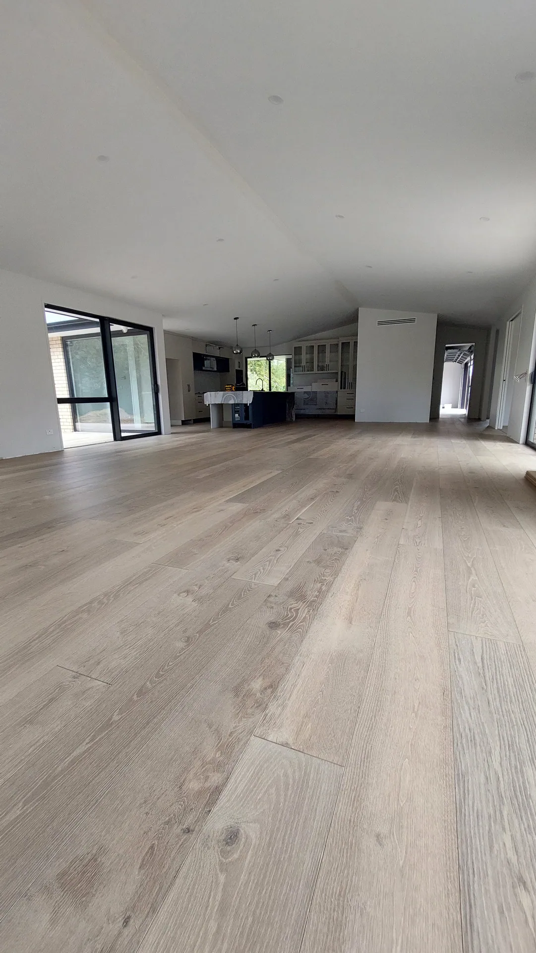 A wide open-plan living area with pale oak boards running toward black-framed windows and a kitchen.