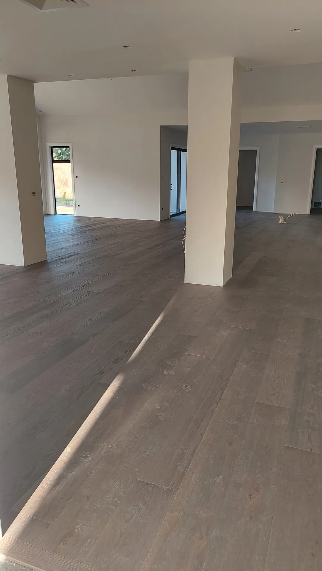 An empty open-plan space with square columns and a dark smoked oak floor crossed by a streak of sunlight.