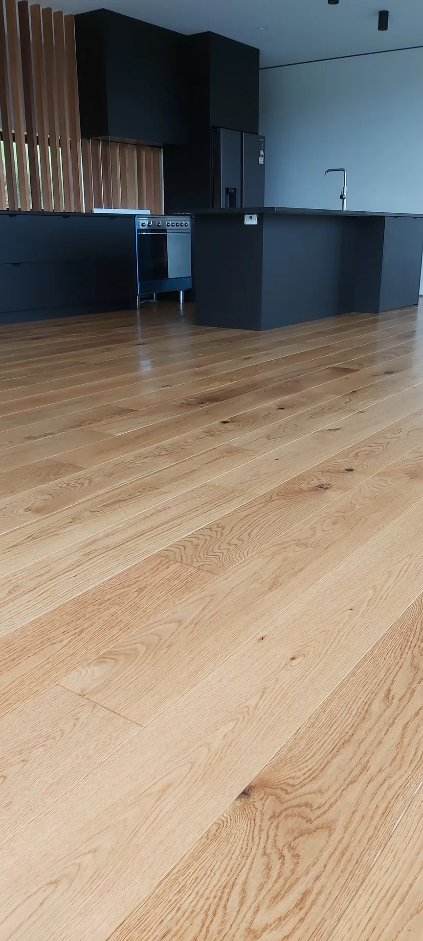 Low-angle view along wide light oak floorboards towards a modern black kitchen with timber battens.